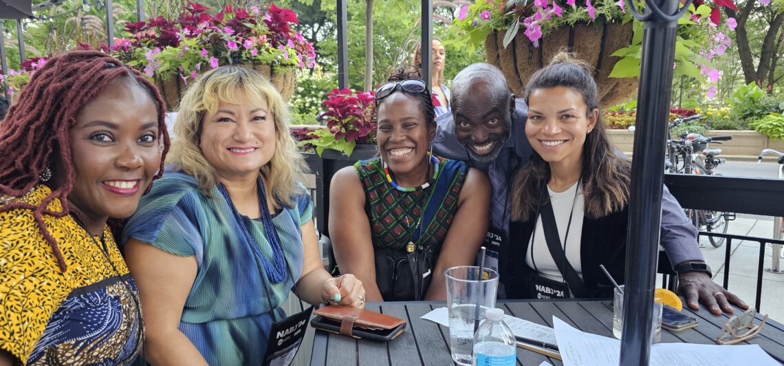 A group of five people smiling and sitting together at an outdoor table, surrounded by plants and flowers.