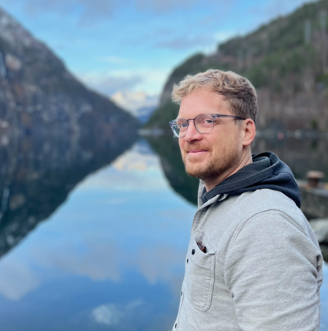 Photo of a fair-haired man wearing glasses and a tan outdoor jacket standing in profile before a waterway and hills.