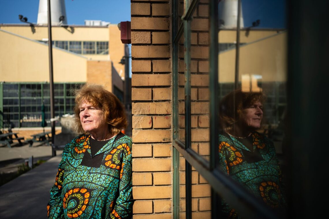 A woman with red hair, wearing a colorful chevron-patterned dress, leans against a brick wall, her reflection visible in a window.