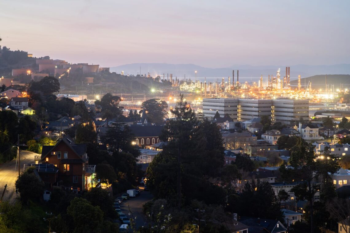 A nighttime view of a coastal industrial area with illuminated buildings and residential houses in the foreground, perfect for city news site stories.