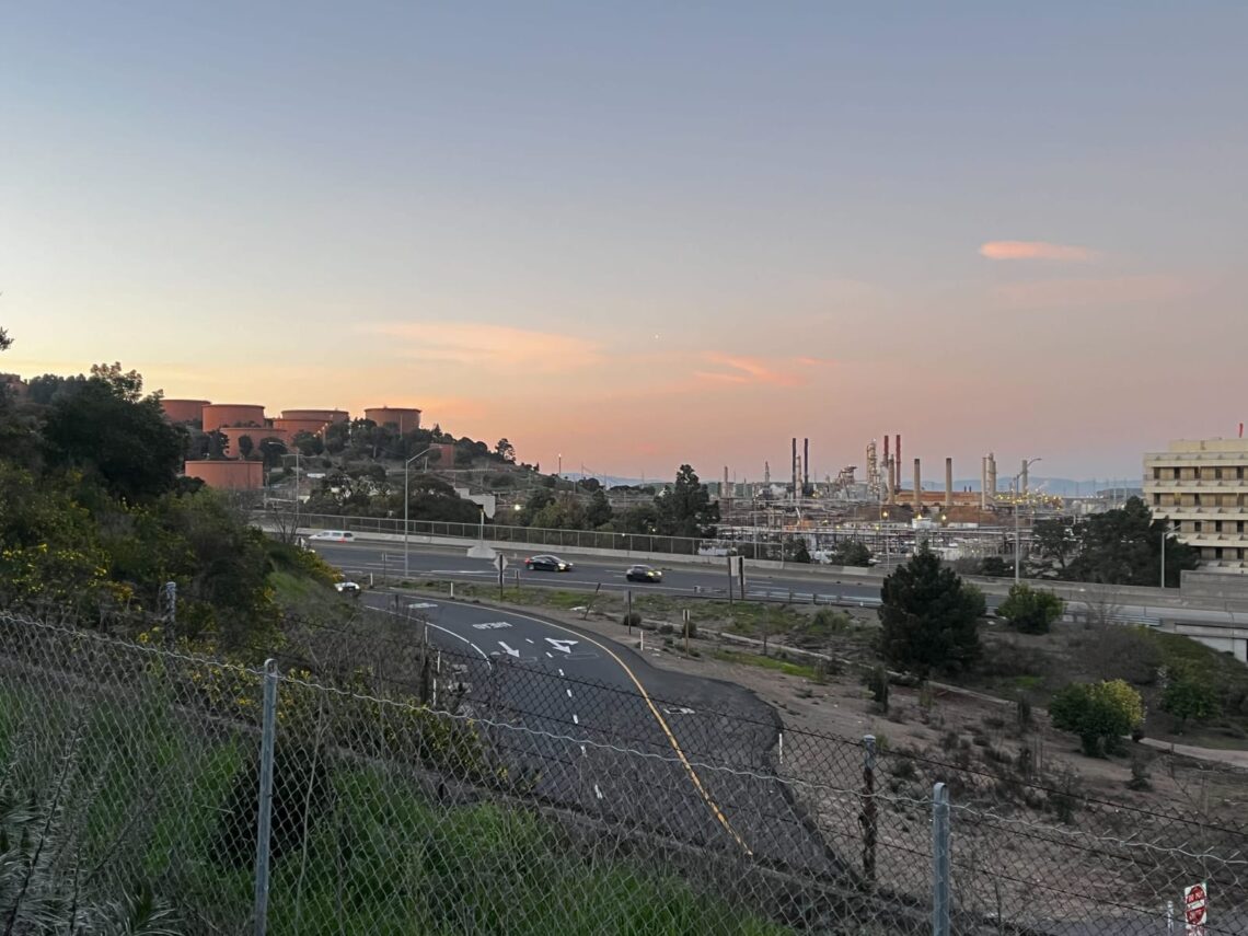 Sunset view of an industrial area with a highway in the foreground and factories with smokestacks in the background, reminiscent of stories you might find on a city news site.