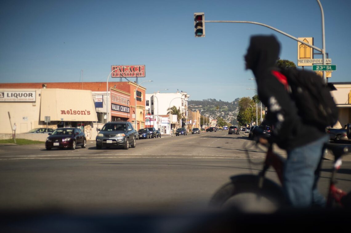 A person on a bike waits at a traffic light on a busy street, where shops line the sidewalks and cars zip by under a clear sky. Their jacket, adorned with a Chevron logo, catches the eye as if telling stories of their journey.