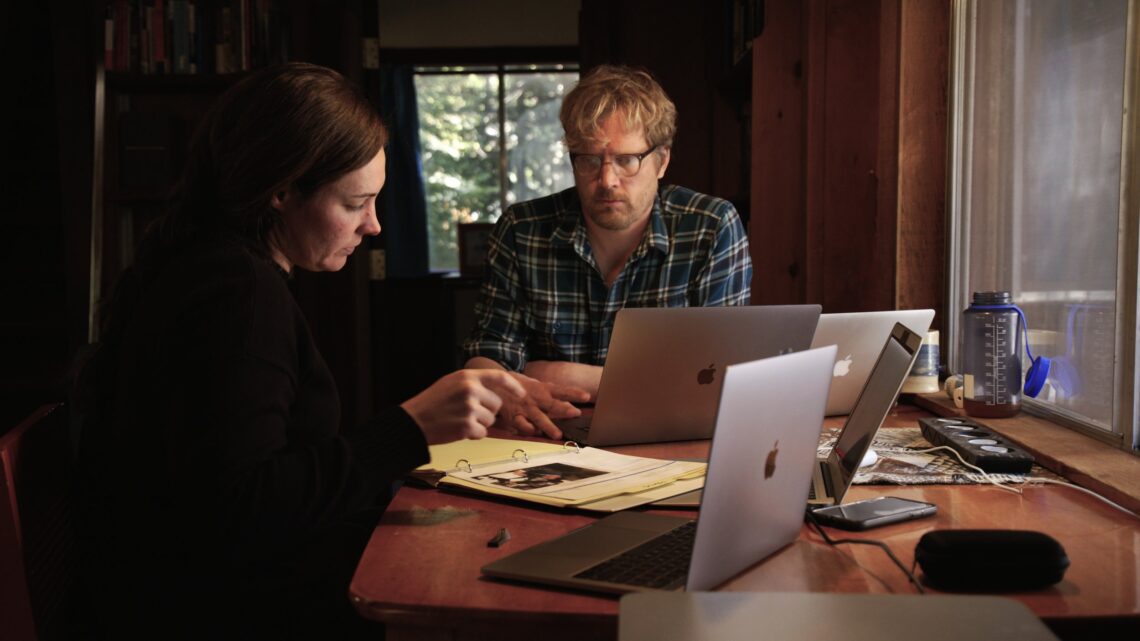 A woman with dark short hair and a man with fair short hair wearing glasses look closely at a computer screen inside a cabin.