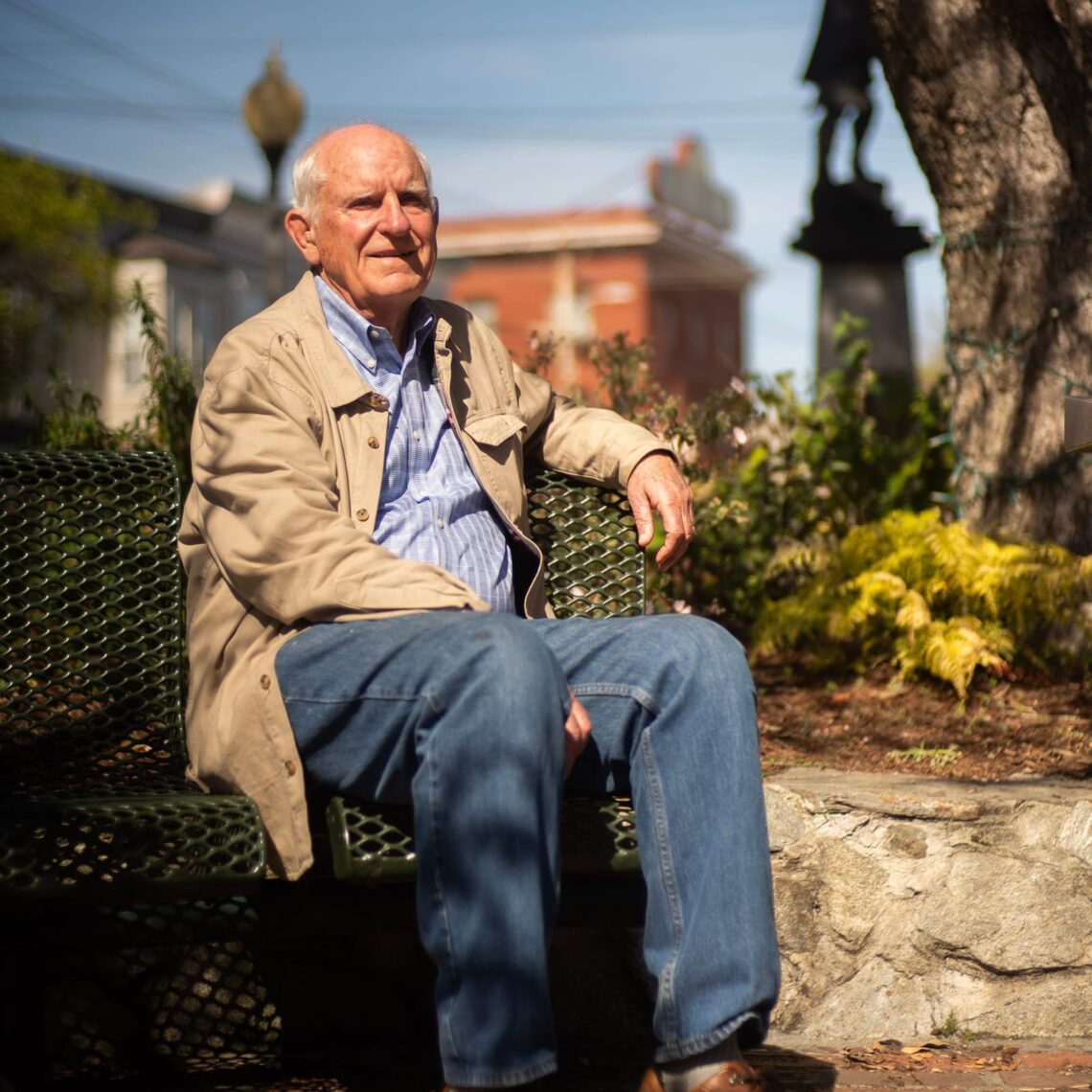 An elderly man in a beige jacket and jeans sits on a park bench, lost in the untold stories of his past, enjoying a sunny day with buildings in the background.