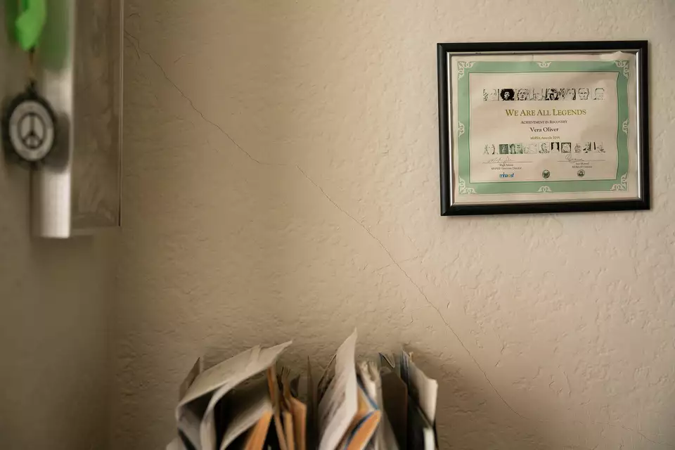 A framed certificate on a wall above a sorted pile of newspapers. On the left a medal with a peace sign hangs by the window.