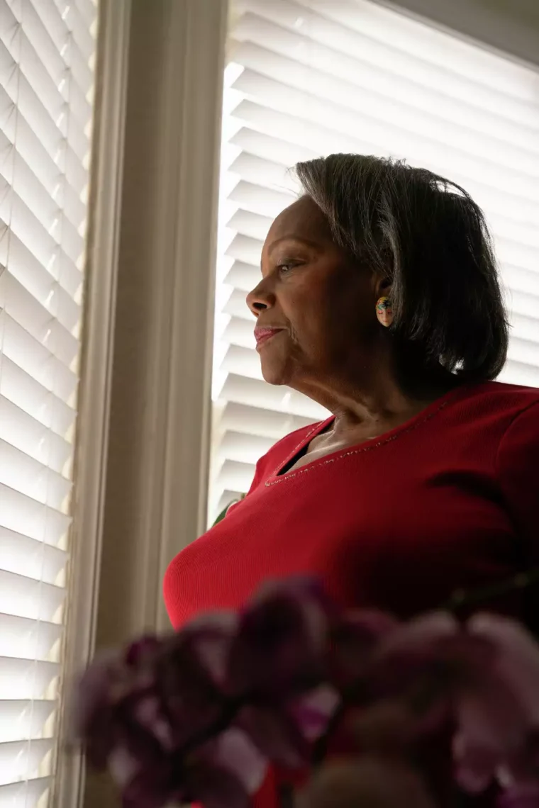 A woman in a red shirt, her eyes serious, peers out of blinds while holding a bouquet of purple flowers. The scene hints at the hidden stories found within the homes of older people.