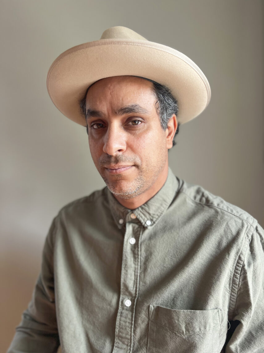 Photo of a man wearing a tan hat and green shirt in front of a beige background.