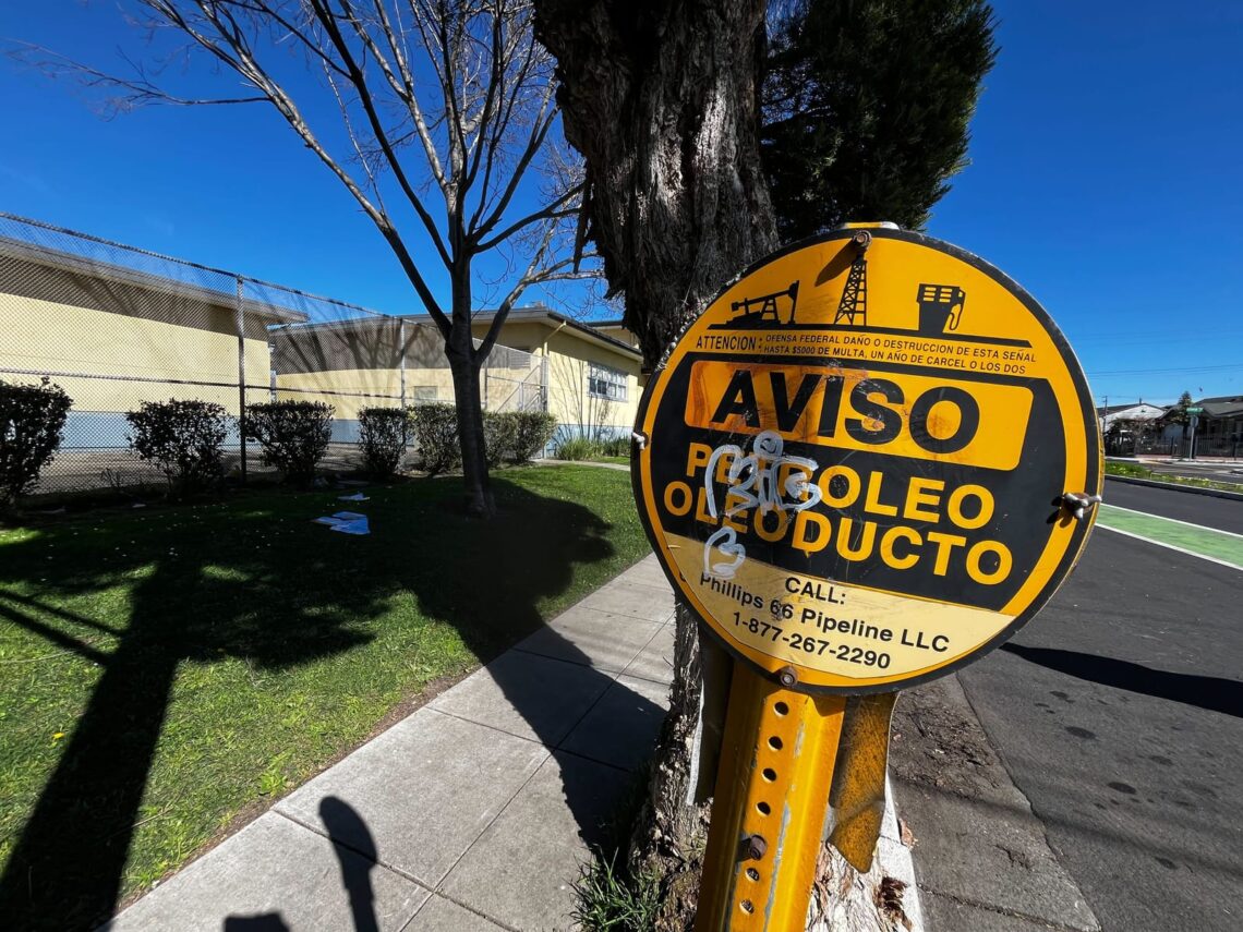 A yellow pipeline warning sign in front of a tree, sidewalk, and building under a clear blue sky tells stories not told in the bustling cityscape.