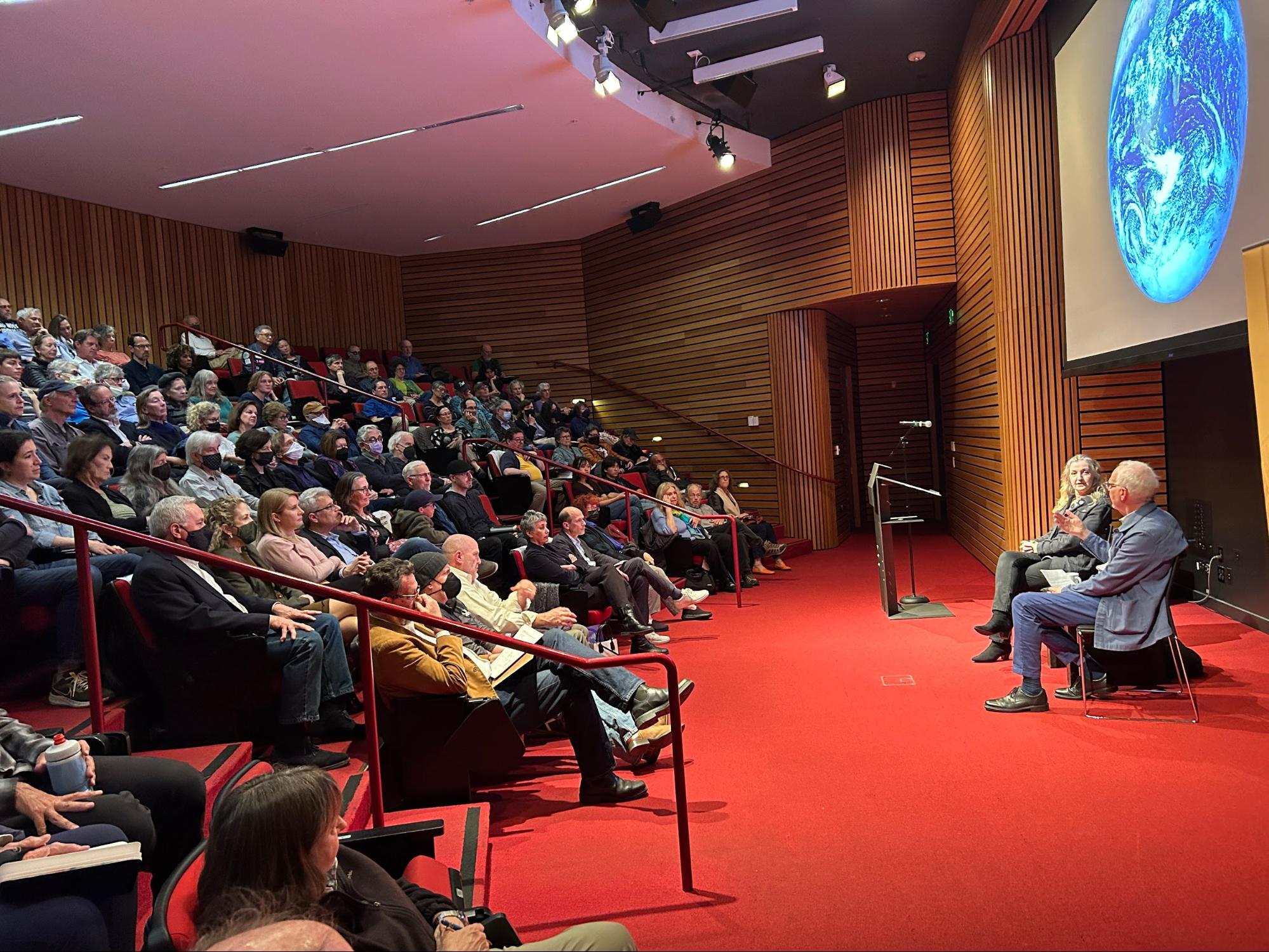 A speaker addresses a full audience in a modern auditorium, with a large image of Earth projected behind them, emphasizing the critical role of climate journalism in today's world.