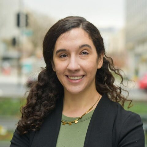 woman with long curly brown hair back in a clip, wearing a green shirt and black jacket. 