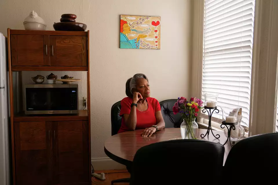 Vera Oliver in a red shirt sitting at a table with flowers, near a window with blinds, and a wooden cabinet filled with keepsakes in the background.