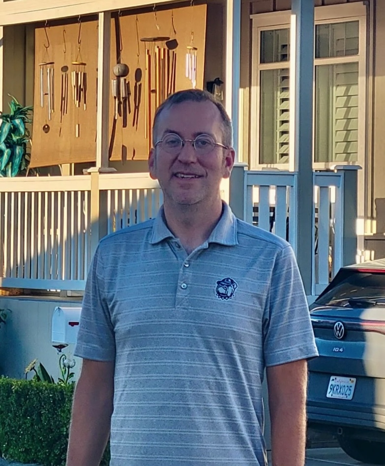 A man in glasses stands smiling in front of a house with a porch, wind chimes, and a parked car in the background, looking like he's ready to embark on his next journalism program.