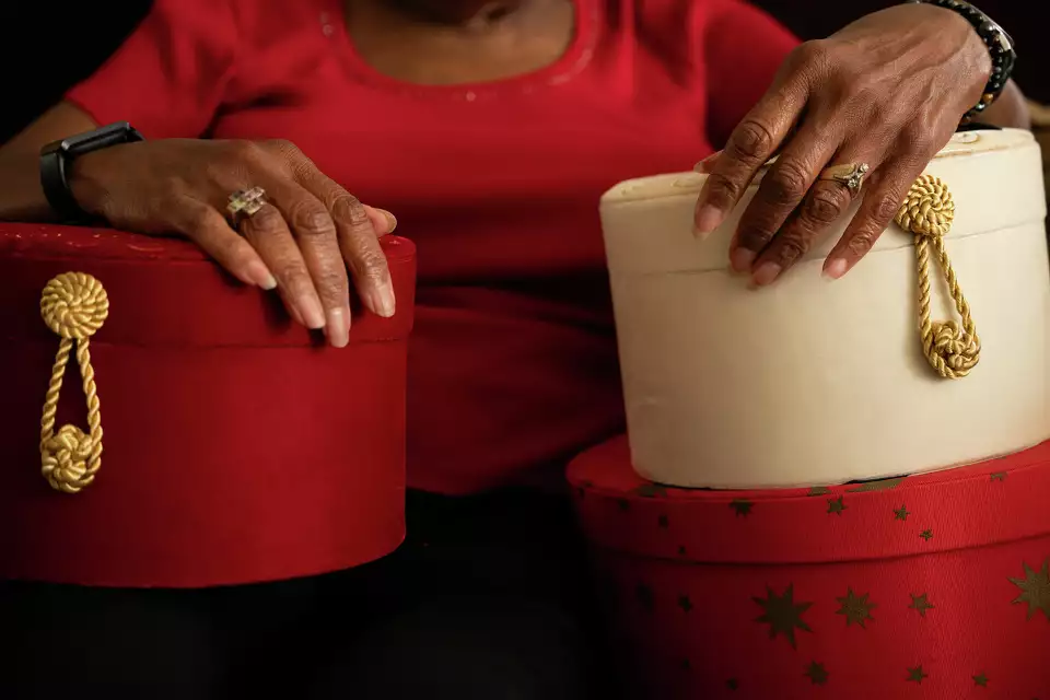 An older person in a red shirt holds two round hat boxes, one red and one white with gold rope handles, reminiscent of homes filled with cherished keepsakes.