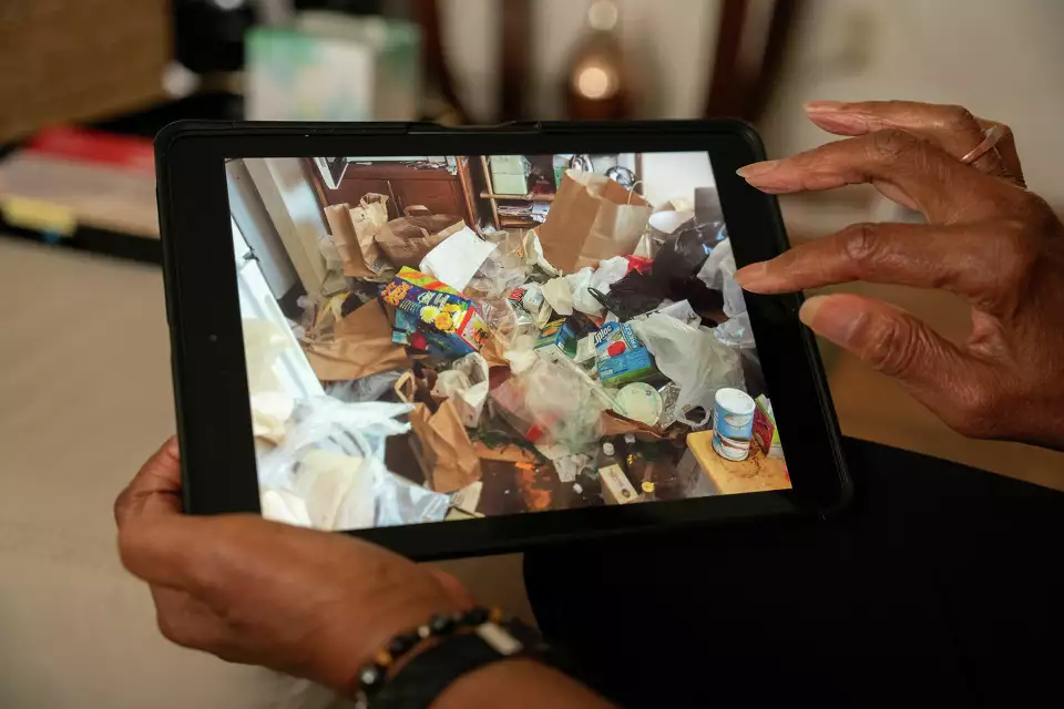 Person holding a tablet displaying an image of a cluttered room filled with various items and shopping bags, highlighting the crisis of chronic hoarding.