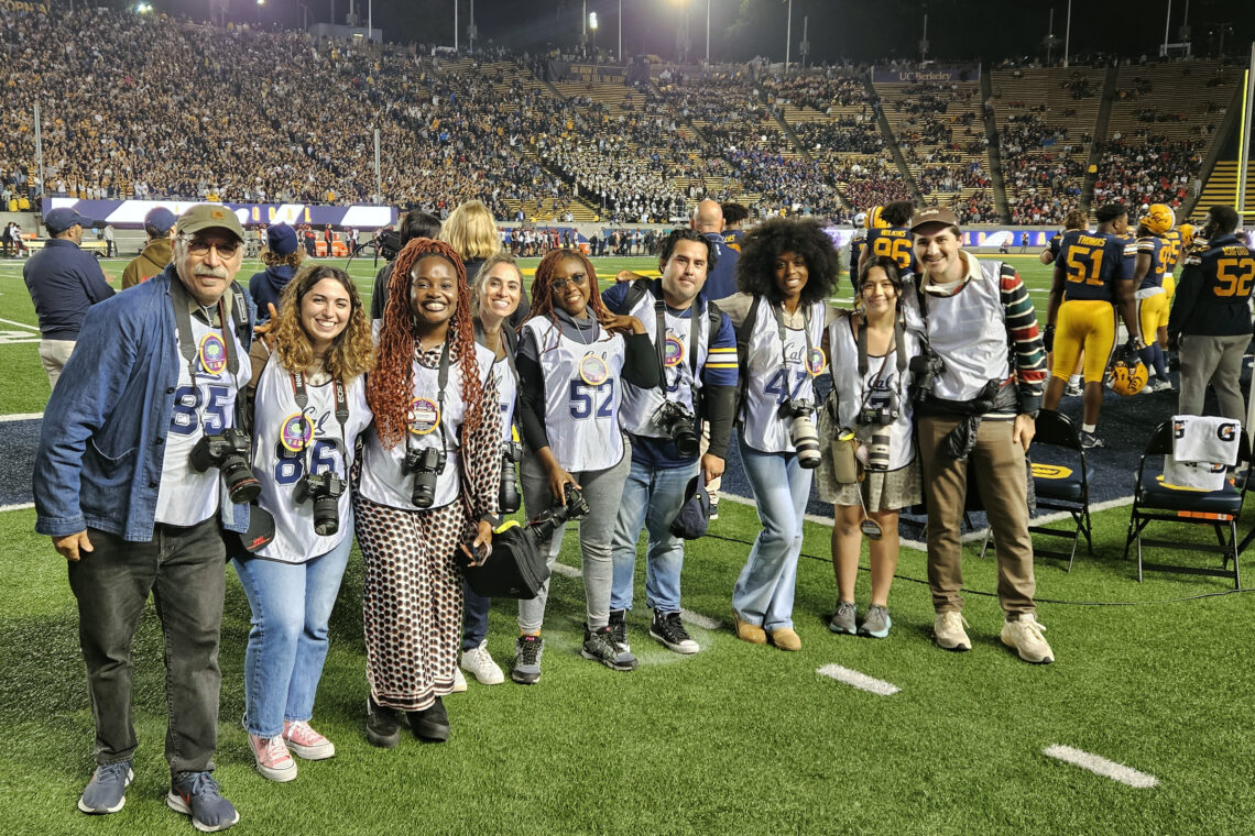 Group of students with camera gear around their necks stand on a football field smiling with their professor.