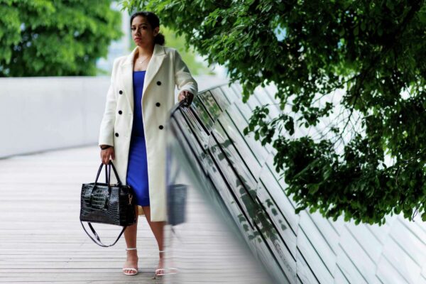 Camile Upchurch poses in Millenium Park in Chicago, she is carrying a black leather purse and wearing a blue dress under a white full-length coat. 