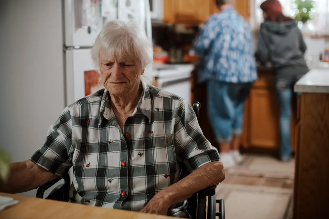 Mary Cooper, an older white woman in a checkered button down shirt sits in a wheelchair at the kitchen table.