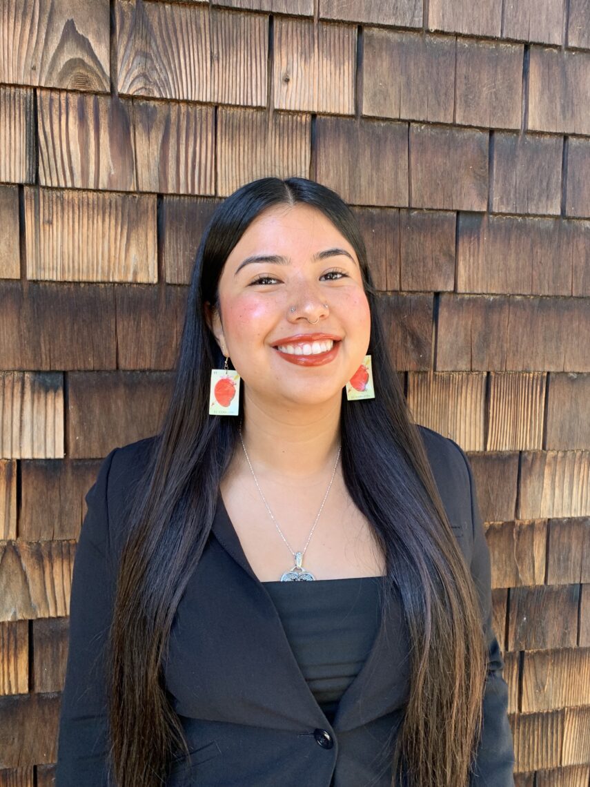 A young woman with long straight dark hair and colorful earrings leans against a building with brown shingles smiling.