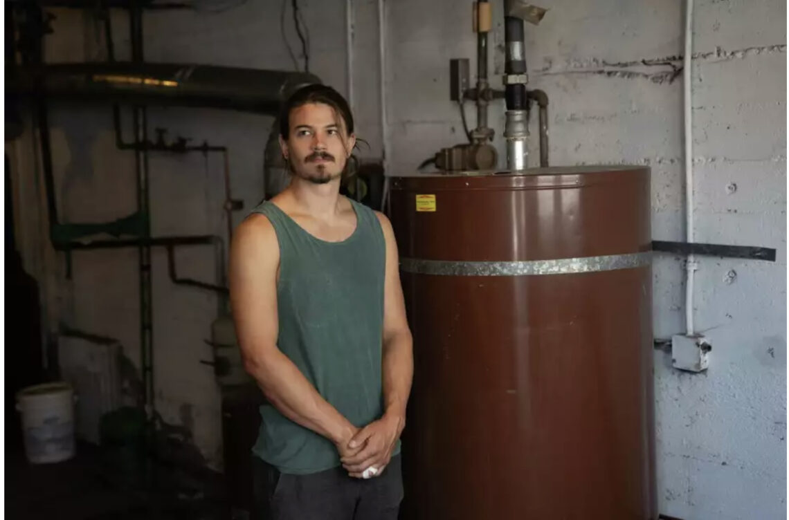 A man with brown hair and mustache wears a green tanktop standing next to a brown water heater.