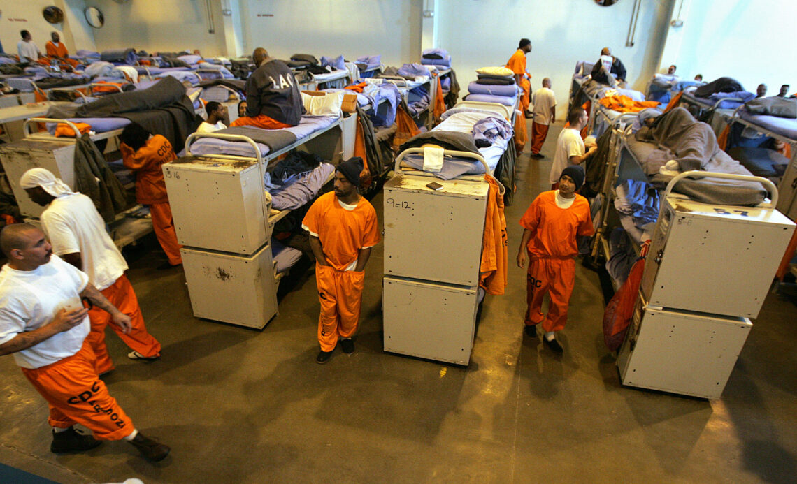 Men in orange CDCR prison uniforms stand and sit in a converted gymnasium filled with metal bunk beds. 