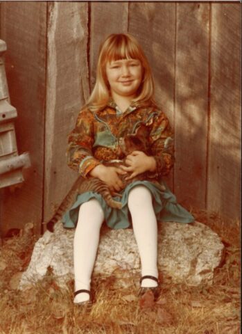 A four-year-old Joslyn Smith sits on a rock in a teal skirt and jacket holding a tabby kitten.