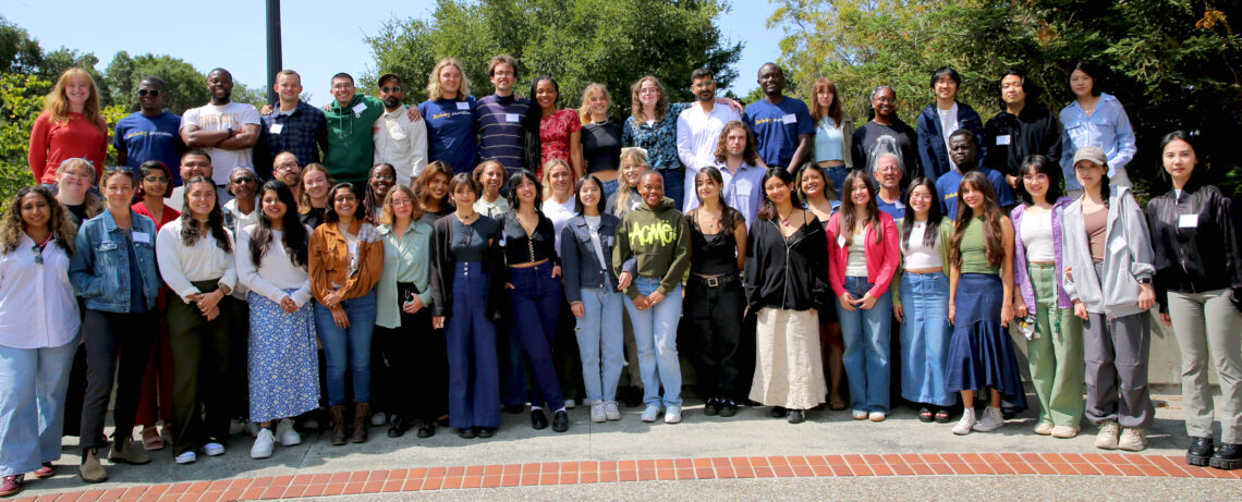 Large group of graduate students standing in rows in front of trees.