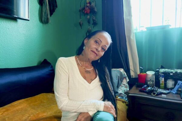 Moonlight Pulido sitting in her home, she is wearing a white sweater and blue pants and looking directly into the camera. Behind her, the teal walls and a dream catcher stand out. 