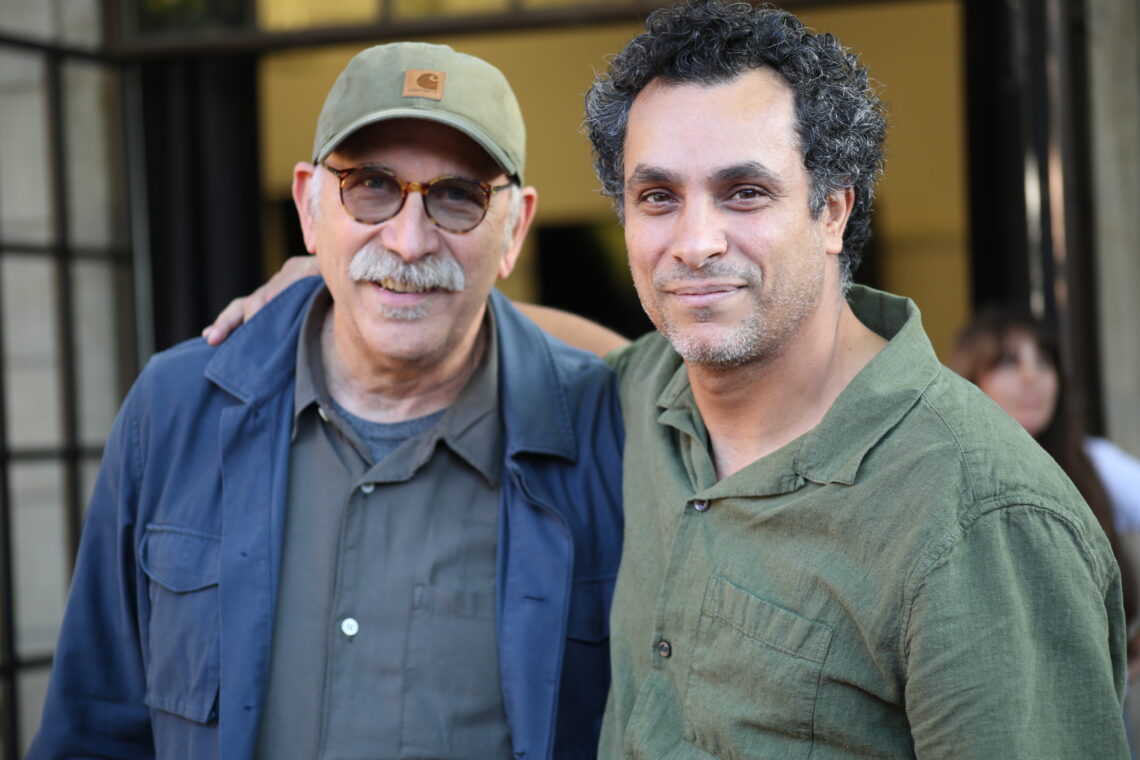 Two men stand close to eachother. One wears glasses and a green baseball hat. The other wears a green shirt and curly dark hair.