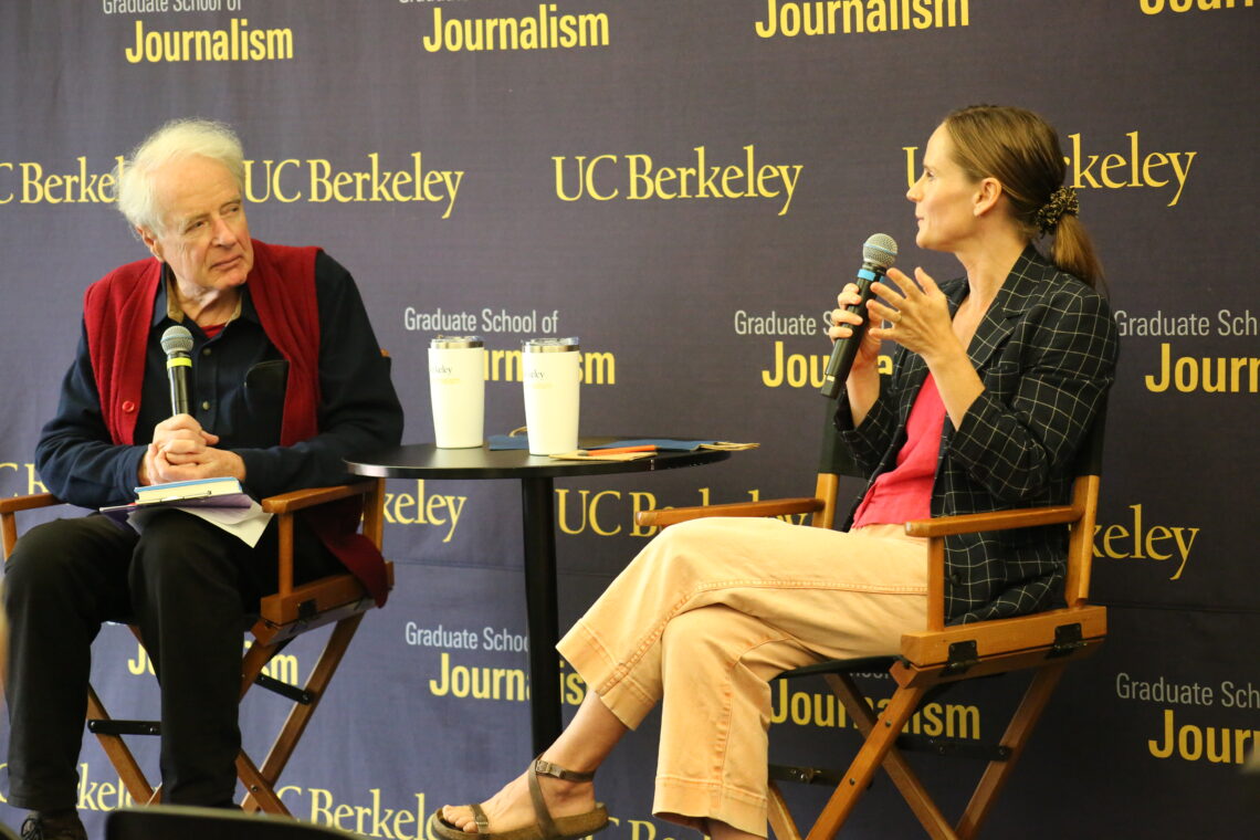 A male professor with white hair sits next to a woman he is interviewing in directors chairs in front of Berkeley Journalism signage.