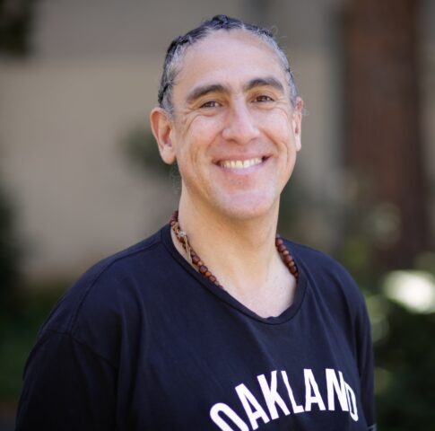 headshot of Professor Andres Cediel smiling with a t-shirt that is blue and reads Oakland. 