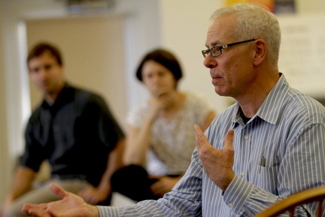 A man wearing a blue and white striped button down shirt gestures with his hands while teaching wearing glasses. Two students appear blurred in the background.