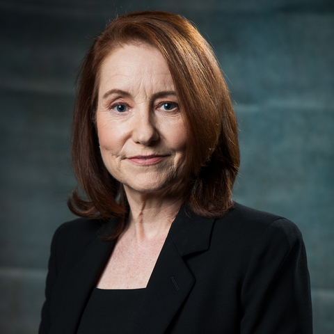 A woman with dark brown red hair in a bob style wears a black suit and looks at the camera.