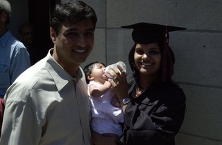 A woman wearing commencement clothes holds a baby next to a man smiling.