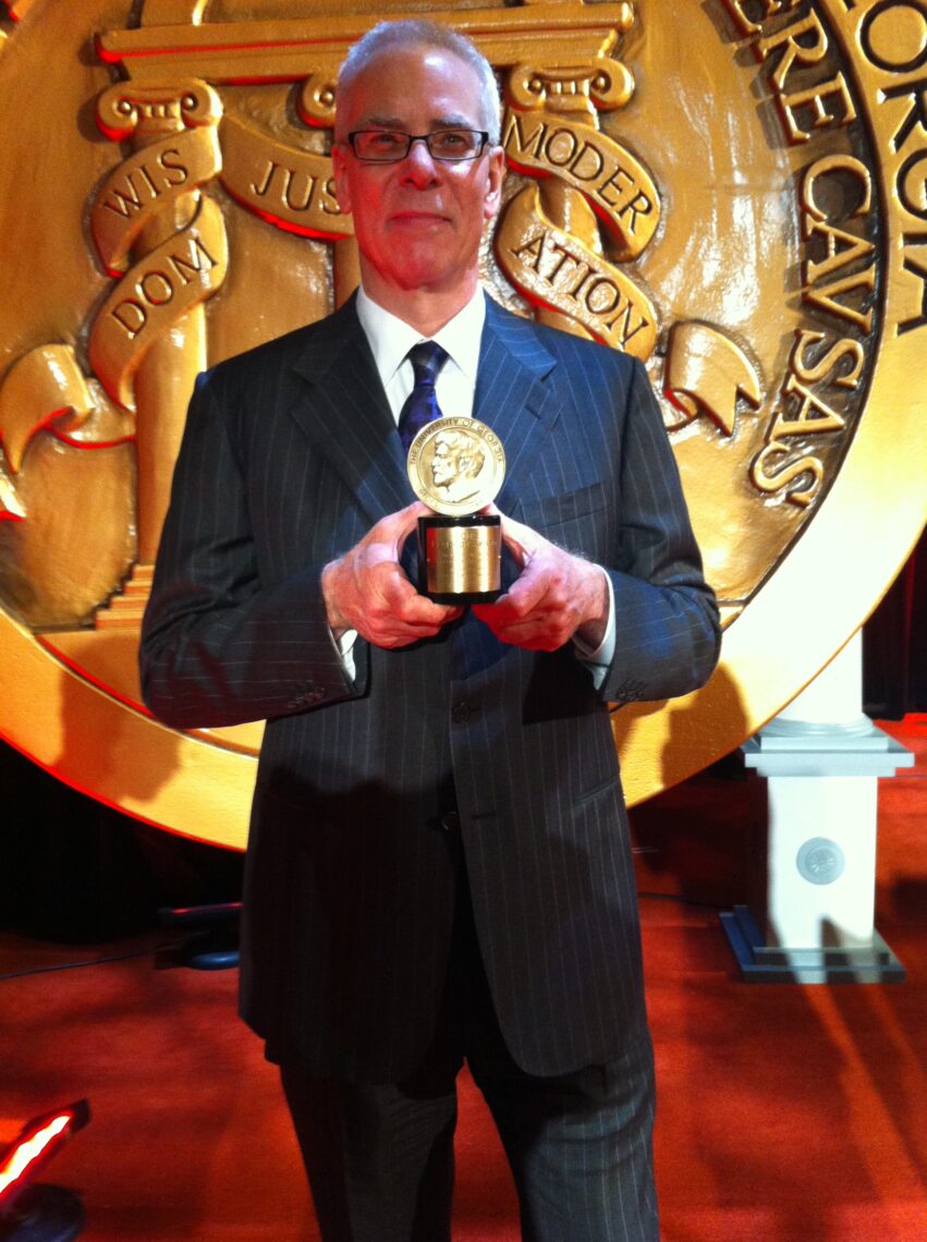 A tall man wearing black glasses wearing a suit holds his Peabody Award in front of signage.