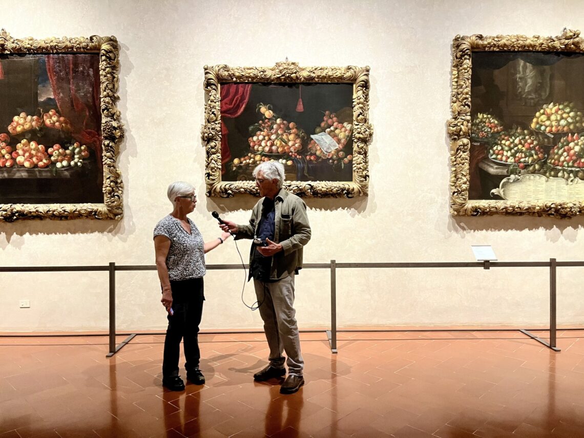 A man wearing a suit interviews a woman in a museum with paintings in the background.