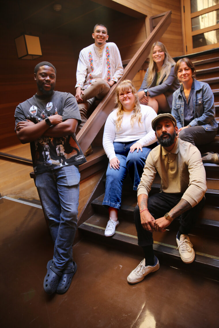 Six students stand or sit on a wooden staircase with filtered skylight light on them.