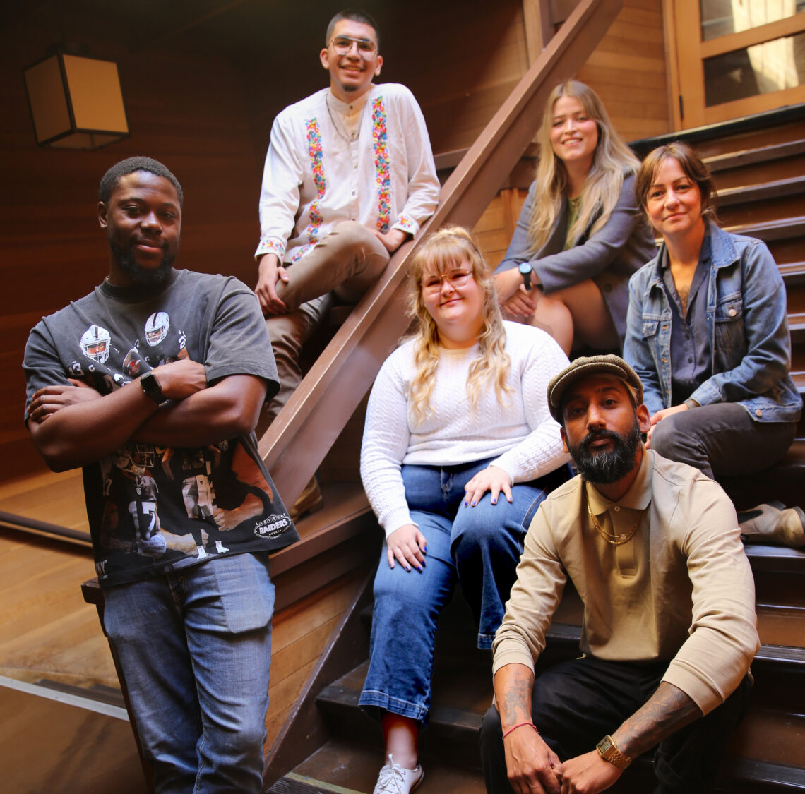 Six students stand or sit on a wooden staircase with filtered skylight light on them.