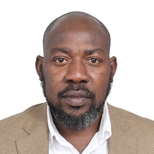 A handsome African man with a beard, mustache and bald head wears a tan suit jacket over a white button down shirt in front of an all white background.