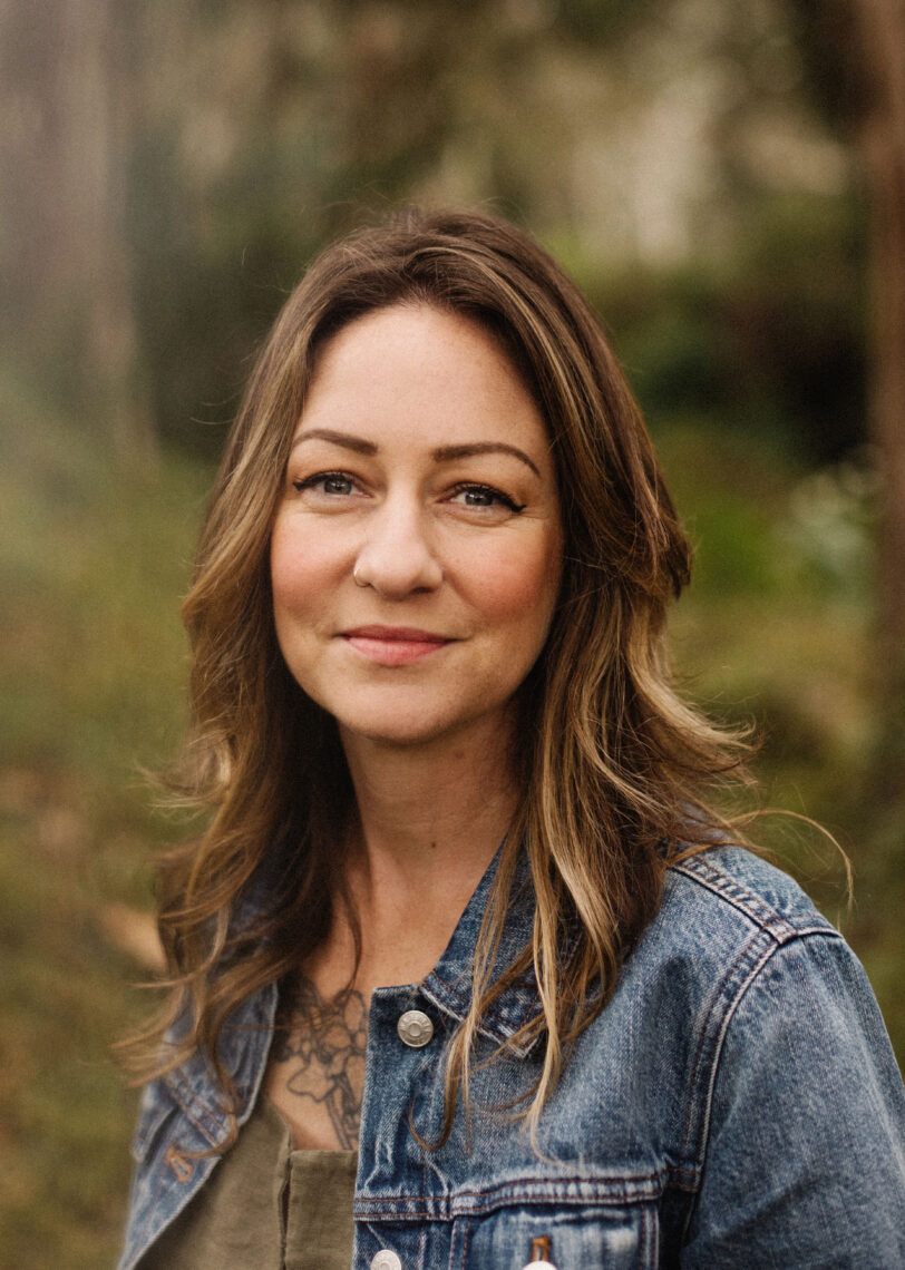 A young journalism graduate student with shoulder length auburn hair wears a denim jacket in front of a green background smiling.