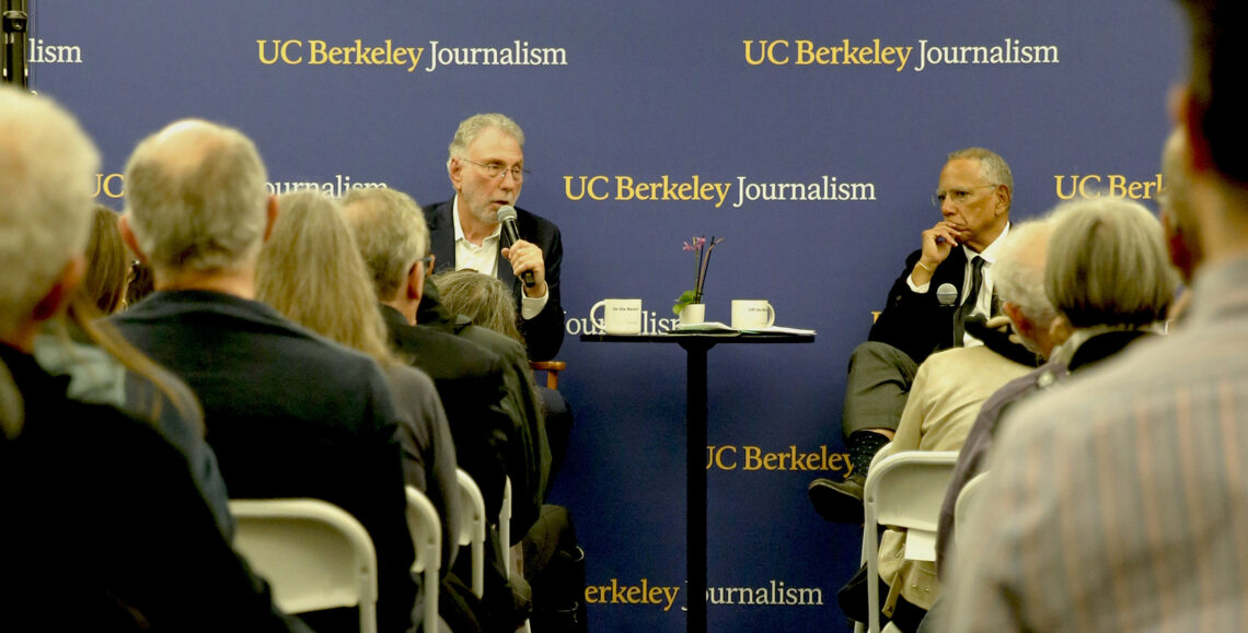 Marty Baron and Dean Baquet talk at North Gate Hall. The image is photographed from the middle aisle. A step and repeat in the background is in Berkeley Blue with the words UC Berkeley Journalism.