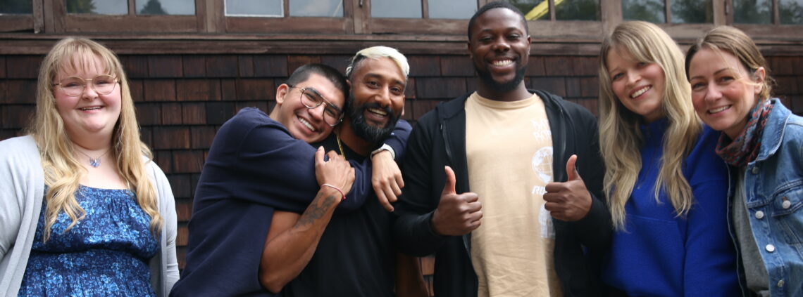 Group of 6 students hugging and looking very happy in front of a shingled building.