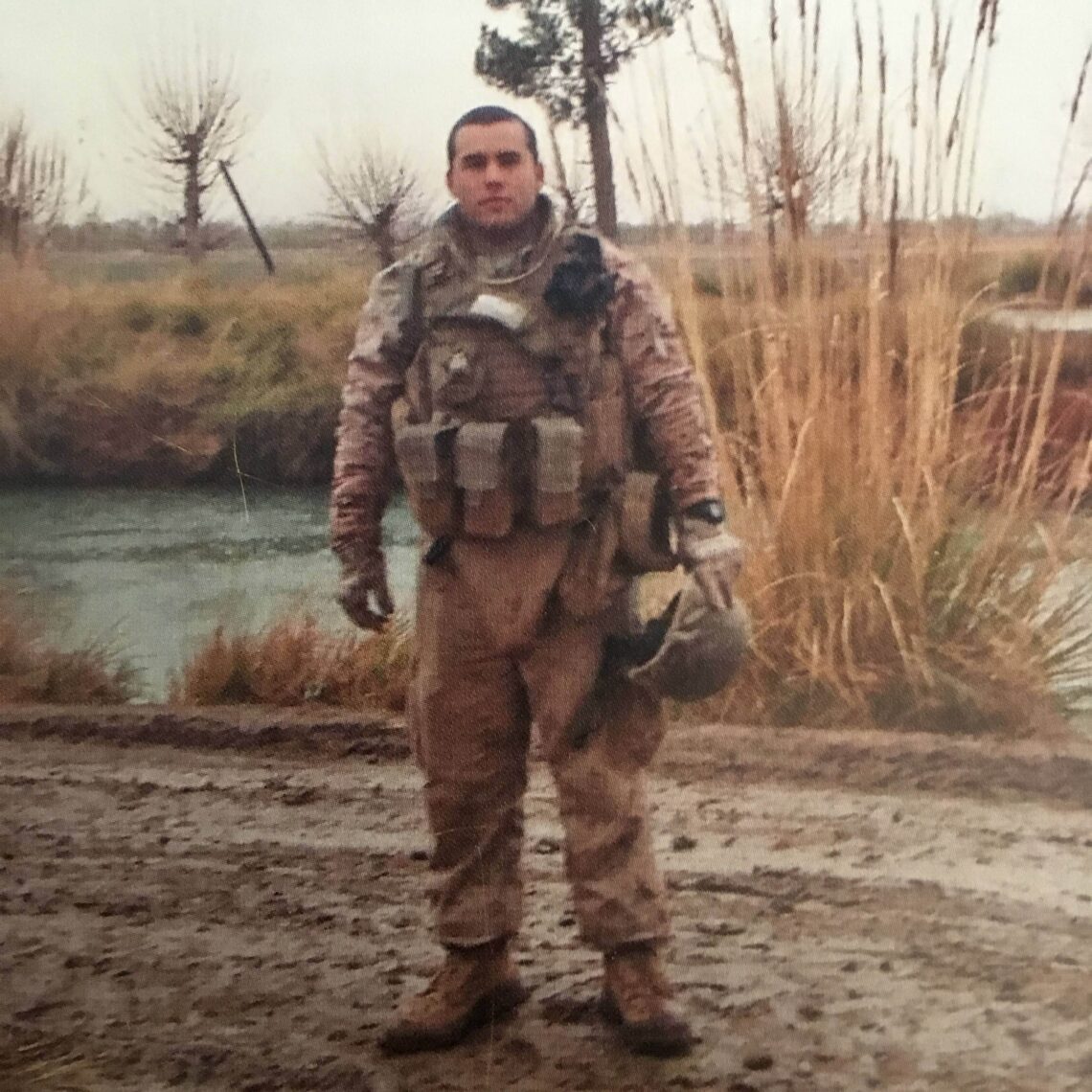 A soldier with dark hair wears military unform in a field.