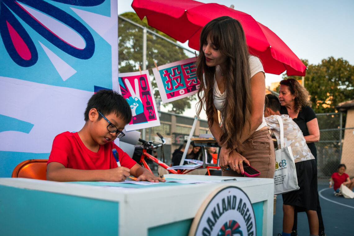 A student looks on as a young person in a red shirt writes something down at a booth in Oakland.