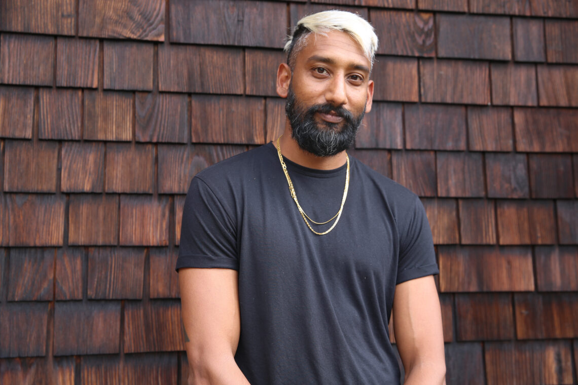 A young graduate student in journalism stands in front of a brown shingled background wearing a black shirt, gold necklace and dark beard with blonde bleached stylishly cut hair smiling.