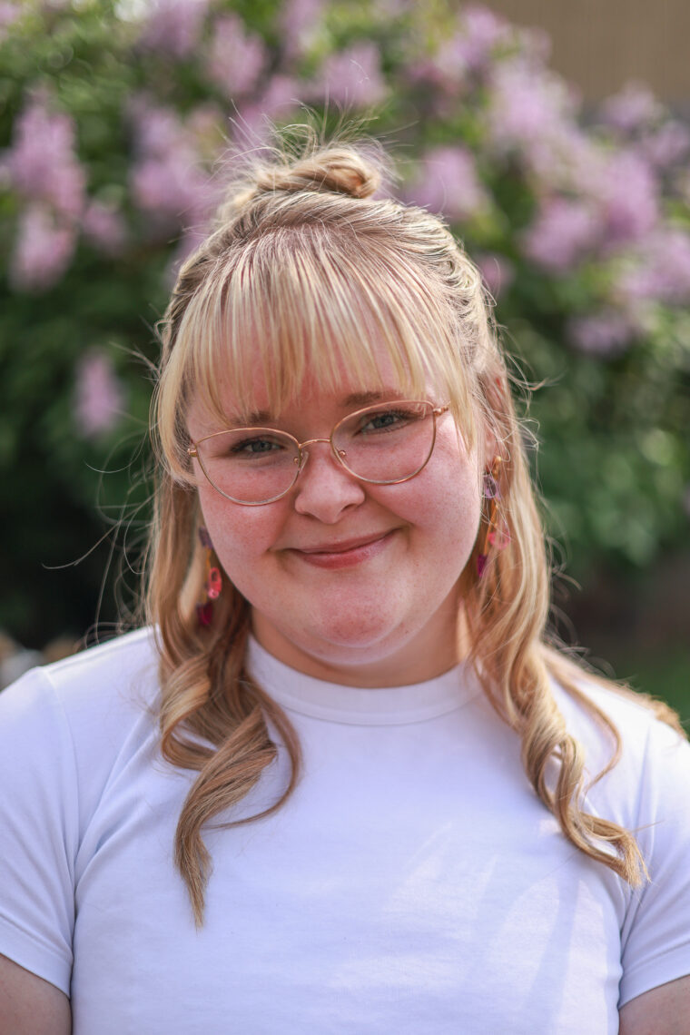 A young blonde woman wears blush color glasses and bangs with her hair up and a white tshirt in front of green and pink flowers.