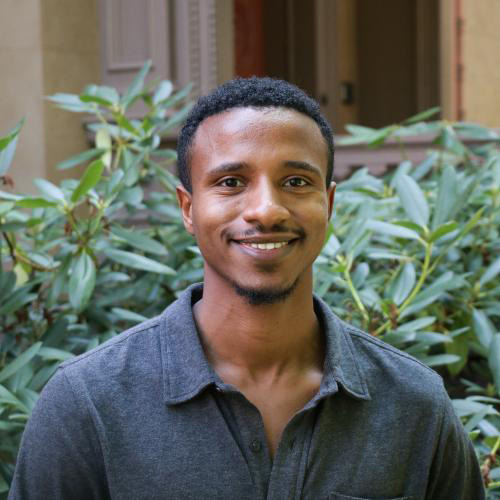 An African American man with short hair wears a dark shirt in front of greenery smiling.