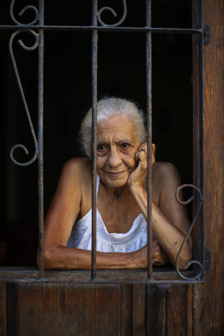 A woman with white hair wears a white nightgown in front of an opening with iron bars and her hand resting against her chin smiling.