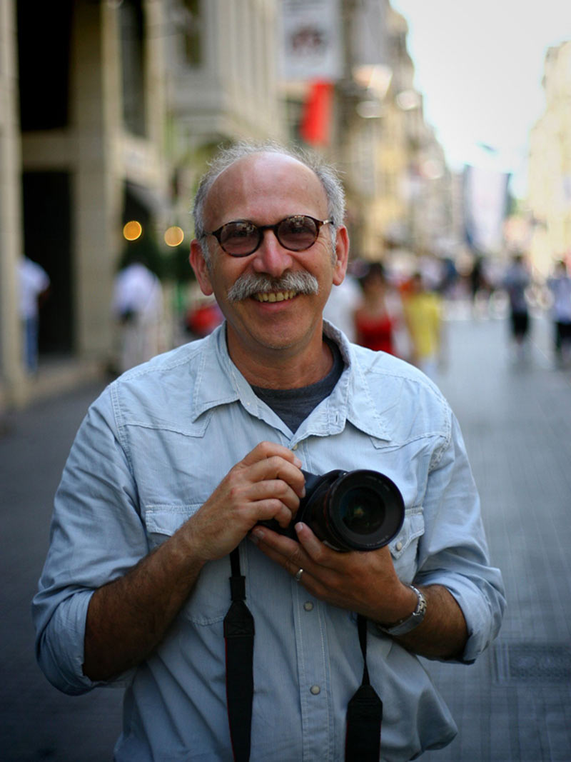 A man wearing glasses and a mustache and white shirt holding a camera on a street.