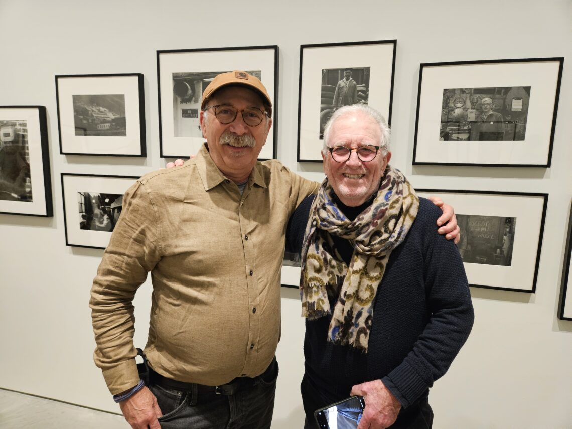 Two older men stand next to oneanother with their arms around eachother's shoulders in front of framed black and white photos at a gallery opening.