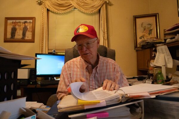 A man with a plaid shirt, glasses, and a red baseball cap sits at a desk and flips through a binder. 